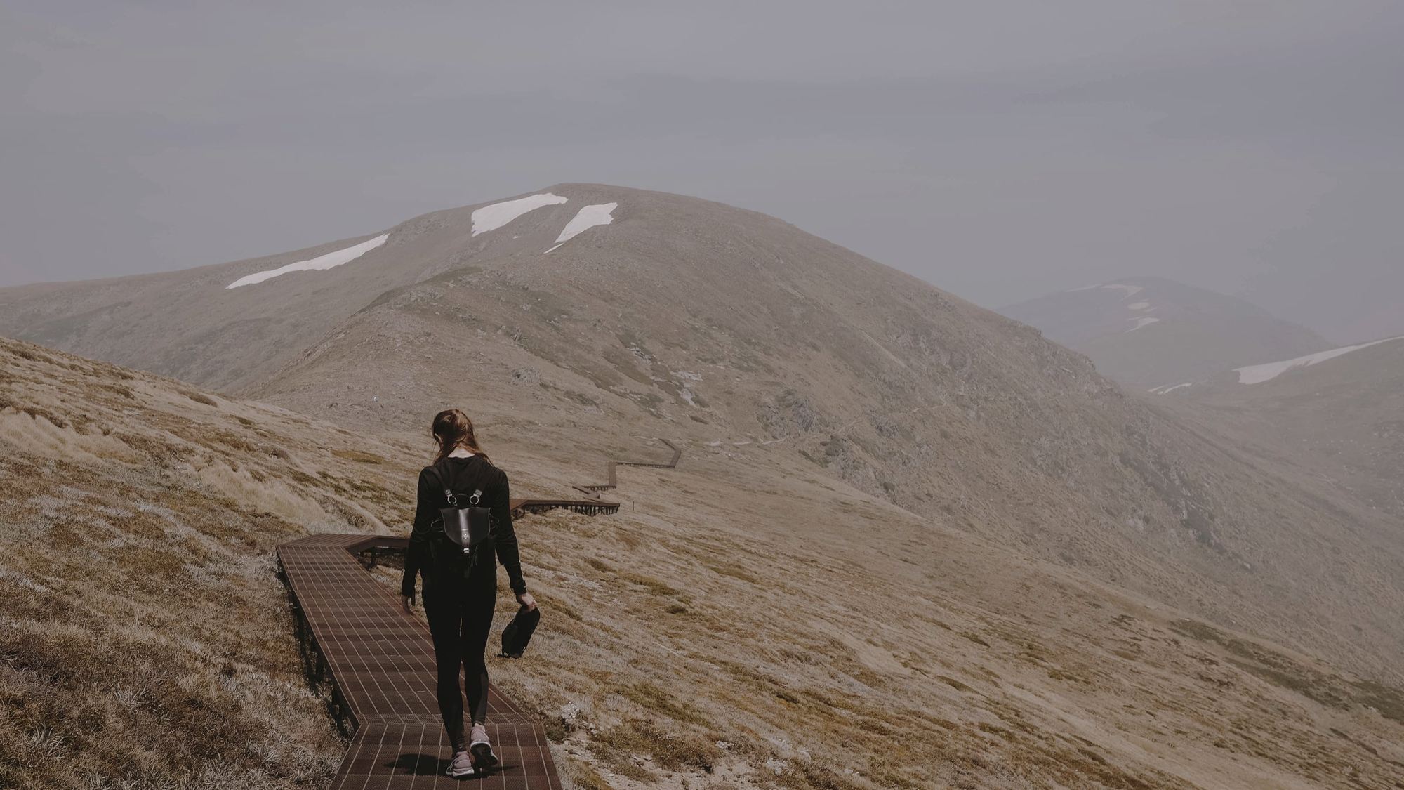 A walker on an alpine boardwalk beneath grey, rounded summits