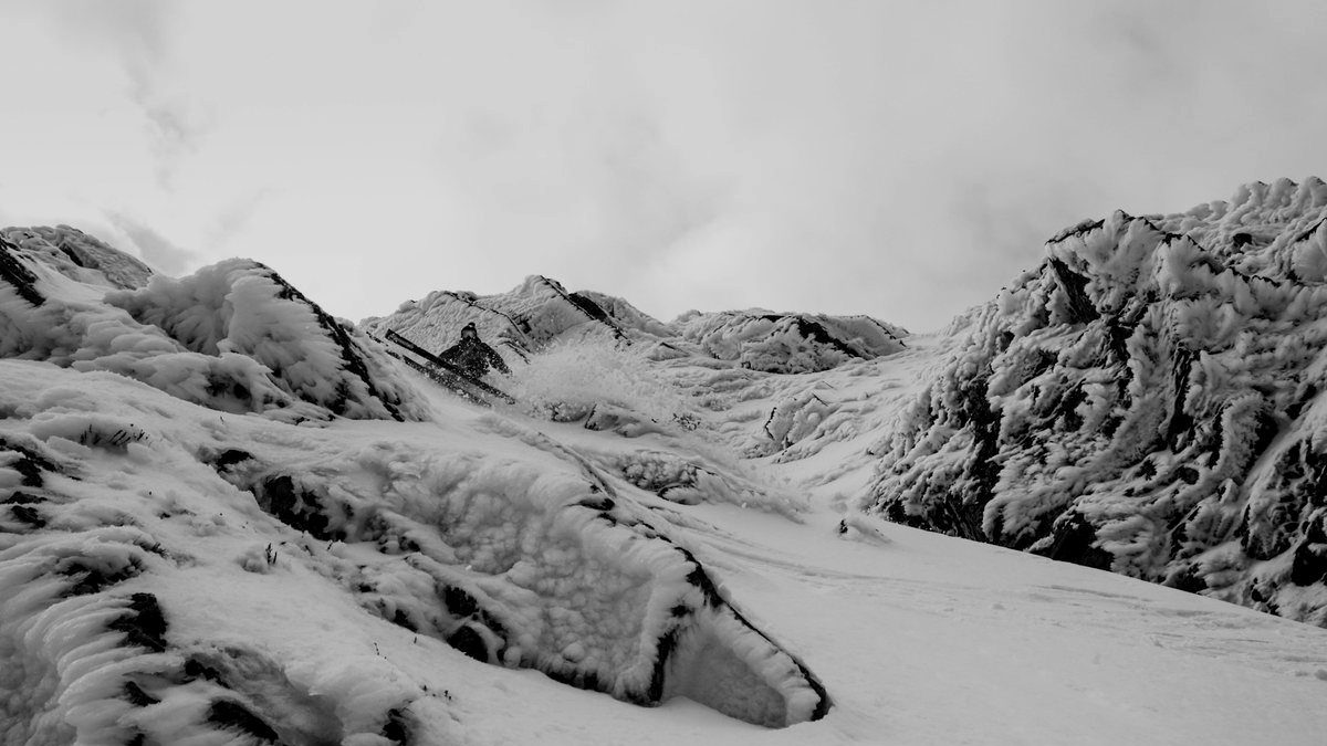 A skier descending a narrow, snowy gully between rime-covered rocks, in black and white
