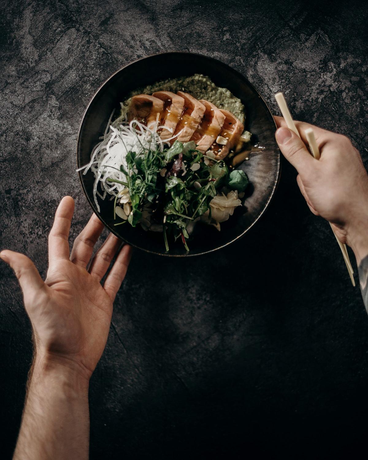 Hands and a dark bowl of vegetables, grains and fish, seen from above