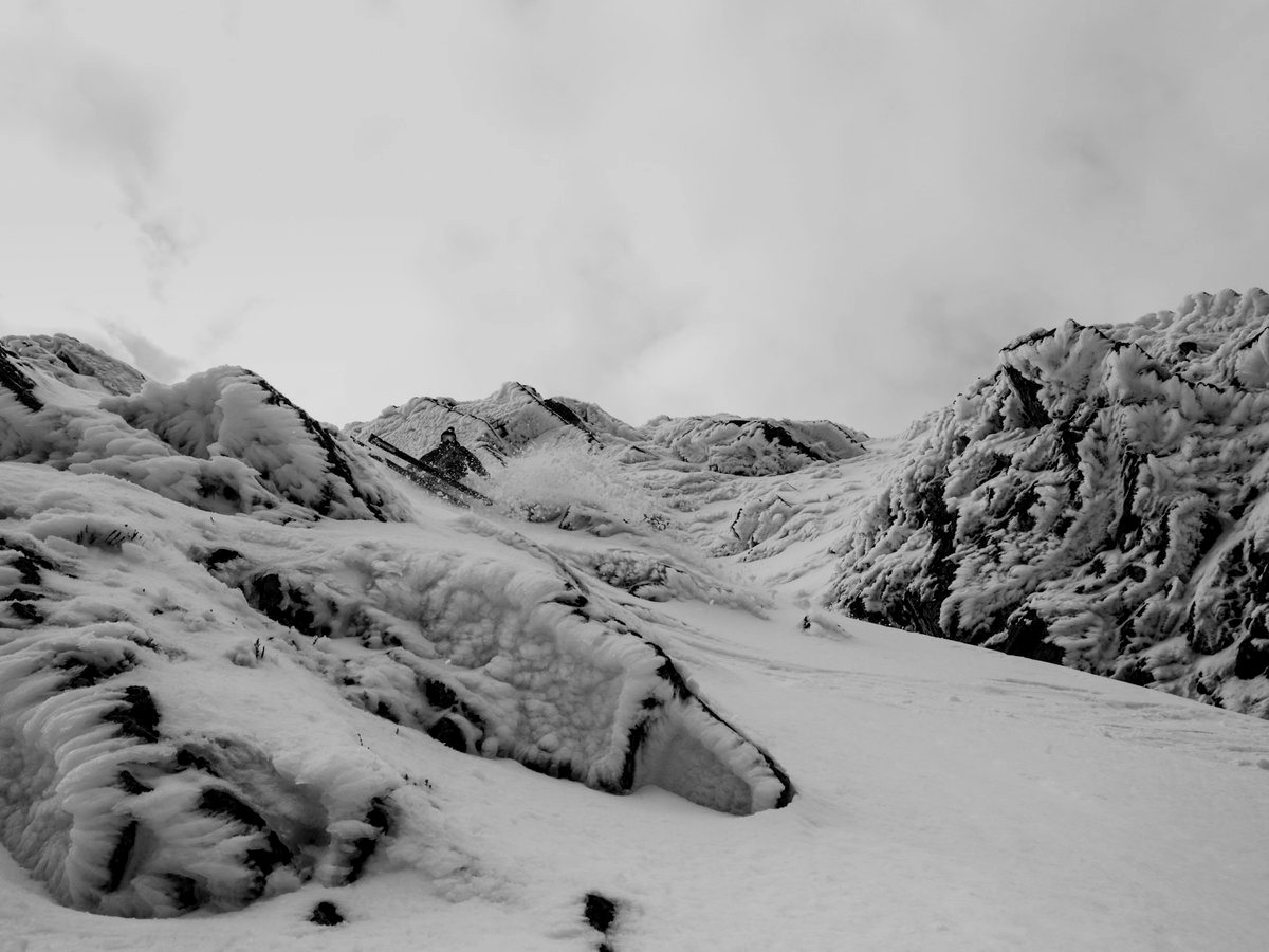 A skier descending a narrow, snowy gully between rime-covered rocks, in black and white